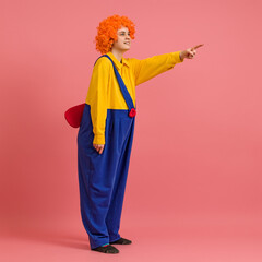 funny clown in a wig and a yellow-blue suit with a propeller on his back stands sideways and points his finger to the side on a colored background