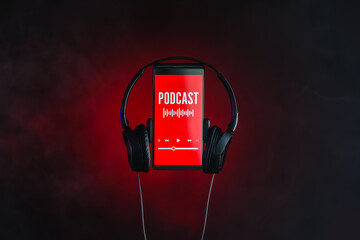 Mobile phone with word PODCAST on screen and headphones on dark color background, closeup