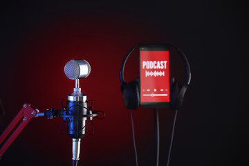 Microphone, mobile phone with word PODCAST on screen and headphones on dark color background, closeup