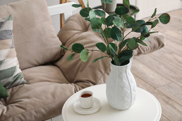 Vase with eucalyptus branches and cup of coffee on table near couch in room