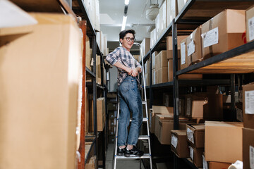 Cheerful female warehouse worker standing on the ladder, next to shelves