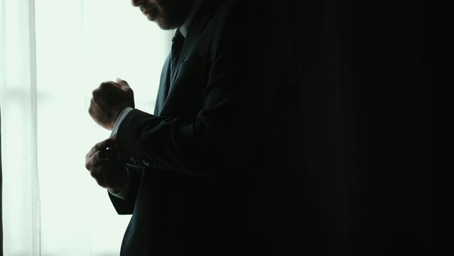 Confident businessman in classic blue suit buttoning or adjust cufflinks near window in hotel room at the morning. Handsome man wearing a nice suit on wedding day.