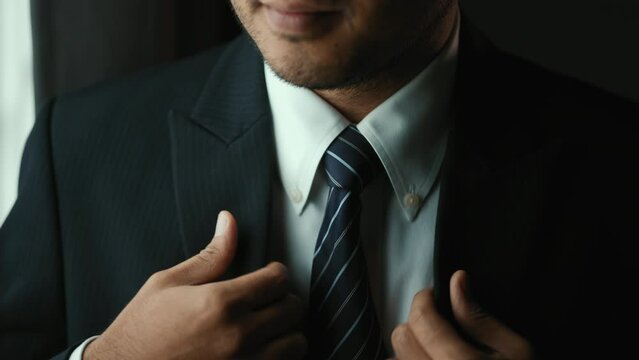 Confident businessman in classic blue suit tying or adjust necktie near window in hotel room at the morning. Handsome man wearing a nice suit on wedding day.