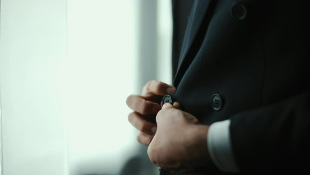 Confident businessman buttoning or adjust classic blue suit near window in hotel room at the morning. Handsome man wearing a nice suit on wedding day.
