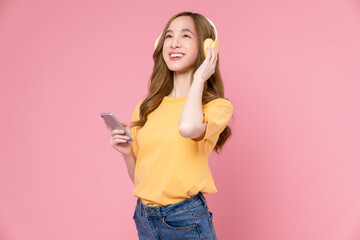 Cheerful young Asian woman in headphones listening to music and enjoy favourite playlist application on smartphone with dancing on pink background.