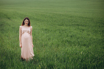 Pregnant woman stand at the field with green grass and embrace her belly. Sunshine summer vibes