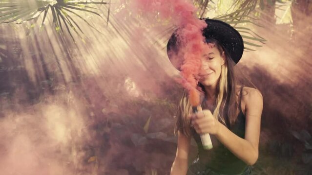 Young beautiful relaxed Caucasian woman teenager moves slowly with smoke bomb in hand enjoys exotic dance or ritual and smiles stands in thickets of tropical plants. Relax, lifestyle