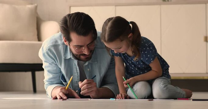 Caring young father in eyeglasses lying on warm floor, teaching small preschool baby daughter drawing pictures in paper album, bonding two generations family involved in domestic hobby activity.