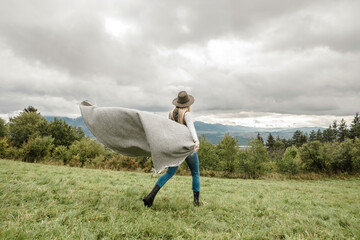 Obraz premium Woman in hat with plaid walks on grass on the top of mountain in camping activity in nature on autumn day, wild forest background. 