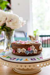 Birthday cake decorated with marzipan flowers on a decorative birthday plate with flowers and a window in the background