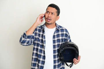 Adult Asian man holding motorcycle helmet and showing confused expression when answering a phone...