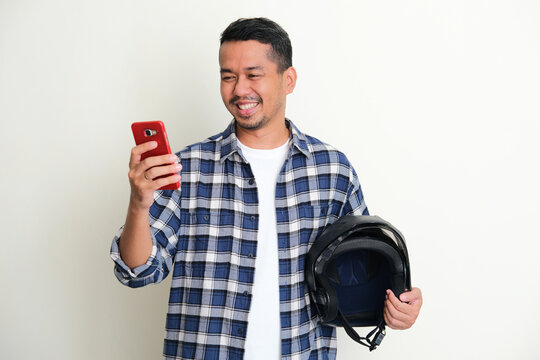 Adult Asian man smiling when looking to his handphone while holding motorcycle helmet
