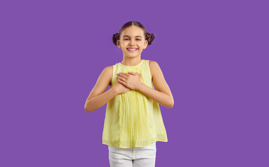 Positive happy grateful preteen girl holding hands on chest isolated on purple background. Kid girl with hairstyle with two bundles happily smiling looking at camera. Concept of joy and gratitude.