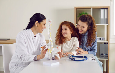 Friendly doctor shows little girl anatomical model of spine standing on table in doctor's office. Joyful mother and her cute redhead little daughter at reception of modern children's clinic.