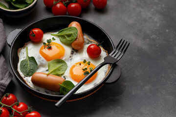 Fried eggs with cherry tomatoes, spinach, sausages in Black pan, top view, dark background.