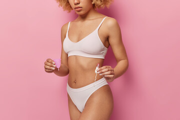 Types of feminine hygiene products. Unknown fit woman holds tampon and menstruation cup things produced for women durinf menstrual periods wears top and panties isolated over pink background - obrazy, fototapety, plakaty