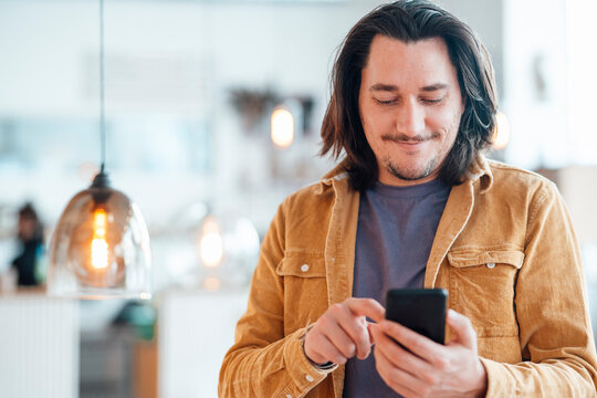 Smiling hipster man with long hair using smart phone at cafe