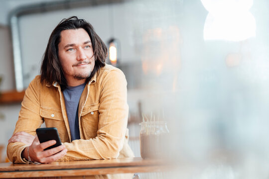 Smiling man with mobile phone leaning on table at cafe - Powered by Adobe