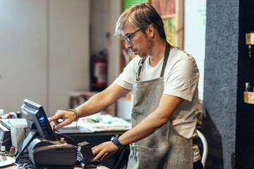 Store owner wearing eyeglasses using cash register at checkout in organic market