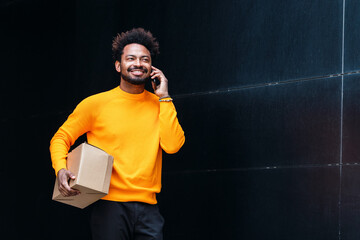 Smiling delivery man holding box talking on mobile phone in front of black wall