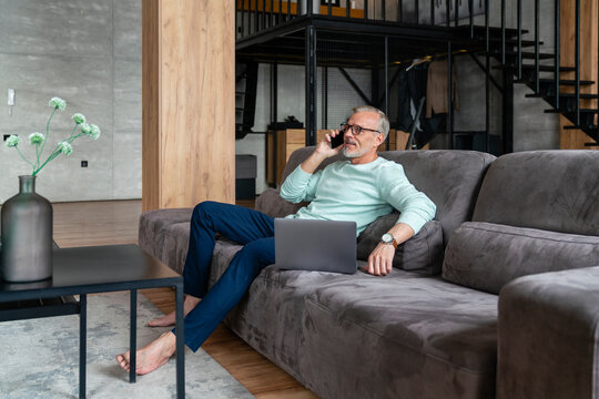 Businessman talking on smart phone sitting with laptop on sofa at home