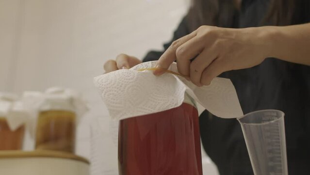 Woman puts tissue cover on glass jar with kombucha ingredients and closes it with elastic rubber band.