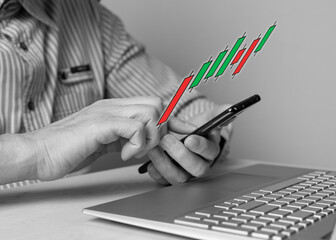 Man with phone in hands using candlestick graph for tracking price movement. Stock market analysis, investment, trade, finance concept. Male sitting at table with laptop. Black and white. photo