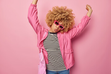 Indoor shot of happy carefree woman has curly hair keeps arms raised up moves with rhythm of music feels joyful wears trendy sunglasses striped jumper and jacket feels glad and full of energy.