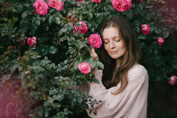 Young woman wearing pink dress enjoying fragrance of blooming roses in a spring garden.