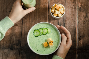 Female hands holding a plate cucumber gazpacho. on a wooden table, top view. Healthy vegan food. 
