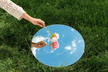nature concept - hand with peony flower and sky reflection in round mirror on summer field