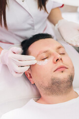 Fototapeta premium Cosmetologist applying anesthetic cream on man face in beauty salon