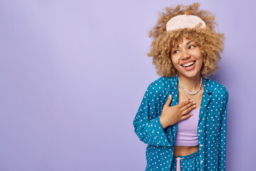 Positive young woman dressed in blue pajama and sleepmask giggles positively looks away keeps hand on chest smiles broadly isolated over purple background empty space for your promotional text