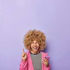 Vertical shot of curly haired woman points index finger above uses mobile phone for chatting online wears casual striped jumper jacket shows blank space for your advertisement isolated on purple wall