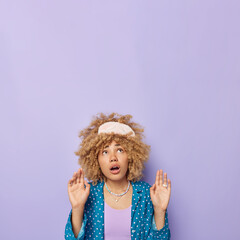 Scared curly haired woman keeps arms up like something falling from above focused overhead with fear wears sleepmask and pajama isolated over purple background empty space to place your promotion