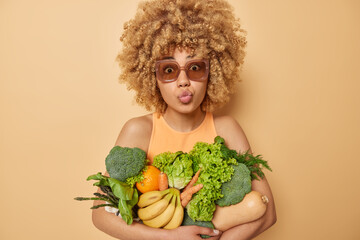 Surprised curly haired young woman keeps lips folded poses with green grocery fullof vitamins eats seasonal vegetables and fruits wears sunglasses and t shirt isolated over beige background.