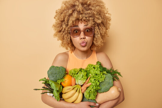 Surprised curly haired young woman keeps lips folded poses with green grocery fullof vitamins eats seasonal vegetables and fruits wears sunglasses and t shirt isolated over beige background.