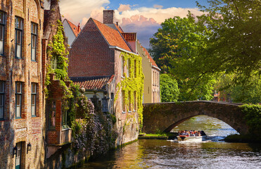 Bruges, Belgium. Ancient medieval stone houses and bridge over canal. Old Stone Bridge between streets. Sunny summer day. Evening sunset with warm lighting. Travel boat with tourists on the river.