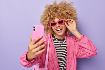 Pretty happy woman with curly hair clicks selfie via smartphone smiles broadly wears trendy...