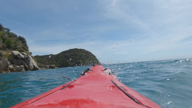 Red sea kayak splashing water towards the camera while crossing over waves in the deep blue sea on a sunny day close to the coastline of Abel Tasman National Park