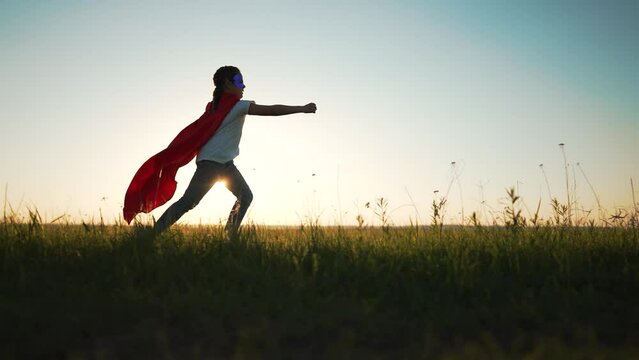 Happy cheerful girl is playing in park. Concept of an active lifestyle. Child in park dressed as superhero. Girl runs through the green grass in the park at sunset. Smiling girl in a superhero mask