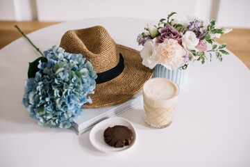 Delicious fresh morning cappuccino coffee,  chocolate cookie, book, blue vase with eustoma flowers, beach hat and blue hydrangea flower on the white table background in a cafe