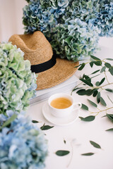Beautiful blossoming blue hydrangea flowers, beach summer hat, eucalyptus and freshly brewed cup of hot espresso coffee with beautiful crema on the white table background, top view, flat lay