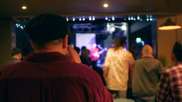 A man watching a band playing on a stage in a music pub club. The man is drinking in the audience