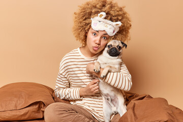 Indoor shot of lovely woman dresssed in pajama embraces favorite pet with love sits in bed keeps lips folded has romantic expression wears sleepmask isolated over beige background. People and animals