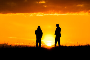 Fototapeta na wymiar Two silhouettes of men talking at sunset or sunrise with dramatic sky and clouds.Dialogue and meeting two people on the horizon and skyline.Outlines of people in the sunlight.Golden hour with friends