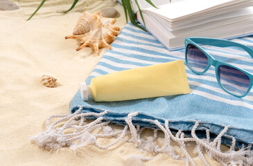 Sunscreen on striped blue towel with book and sunglasses on sandy beach. Background with copy space and visible sand texture.