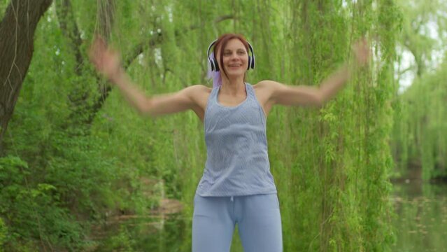 Woman in headphones doing aerobics jumping jacks exercises outdoors. Cheerful female listening the music and doing active sports outside.