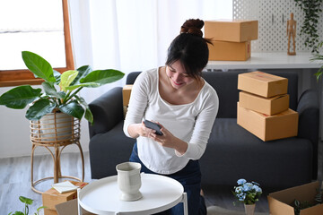Smiling female online seller using smartphone taking photo of product for upload to website online...