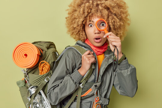 Shocked woman explorer holds magnigying glass over eye cannot believe own eyes explores new place dressed in activewear carries rucksack isolated over green background. Tourism and camping concept - Powered by Adobe